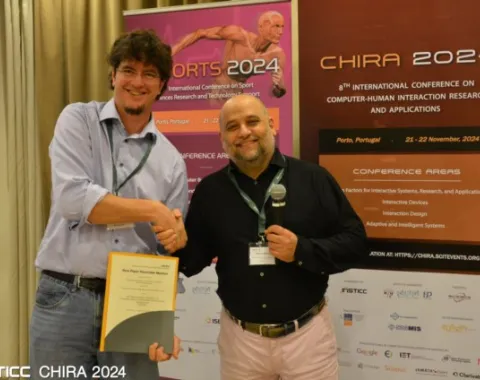 Zwei Männer stehen vor Bannern der CHIRA 2024 Konferenz. Einer hält ein Zertifikat, während der andere ein Mikrofon hält. Beide lächeln und schütteln sich die Hand. Im Hintergrund sind Details zur Konferenz in Porto, Portugal, sichtbar.