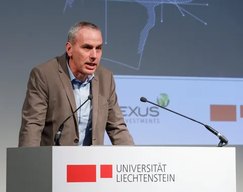 Ein Redner steht an einem Podium mit dem Logo der Universität Liechtenstein. Im Hintergrund sind Logos von PLEXUS Investments und der Universität Liechtenstein sichtbar.