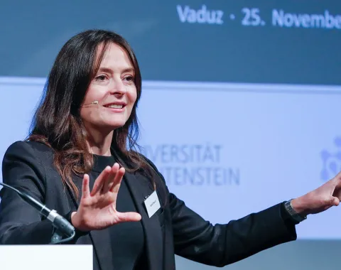 Eine Frau hält einen Vortrag bei einer Konferenz an der Universität Liechtenstein. Sie trägt ein Mikrofon und gestikuliert mit den Händen. Im Hintergrund ist ein Banner mit dem Datum der Veranstaltung und dem Logo der Universität zu sehen.