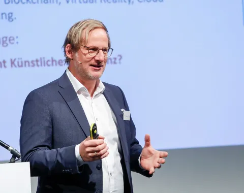 Ein Redner in einem Anzug hält einen Vortrag auf einer Konferenz über Künstliche Intelligenz an der Universität Liechtenstein. Im Hintergrund ist eine Präsentationsfolie zu sehen.