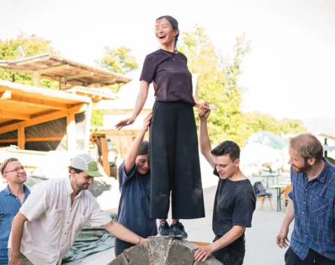 Ein Gruppe von Menschen baut einen selbsttragenden Lehmbogen im Freien. Eine Person steht oben auf dem Bogen, während andere sie stützen. Im Hintergrund sind Bäume und Gebäude zu sehen.