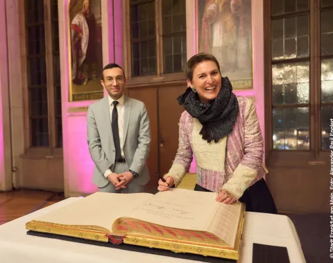 Eine Person in einem eleganten Outfit unterschreibt ein großes Buch auf einem Tisch, während eine andere Person daneben steht. Der Raum ist festlich beleuchtet und im Hintergrund sind Gemälde an der Wand zu sehen.