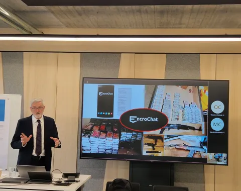 Ein Vortragender steht vor einem Bildschirm, der eine Präsentation über EncroChat zeigt. Der Raum wirkt modern und ist für eine akademische Veranstaltung eingerichtet.