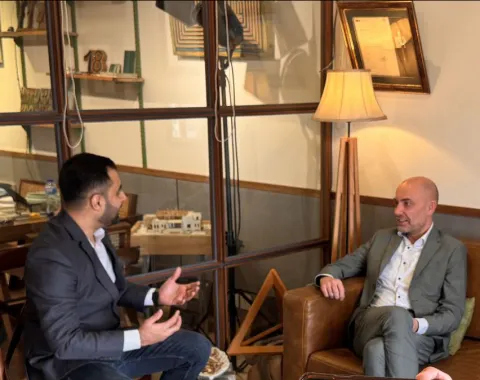 Zwei Männer in einem Gespräch in einem gemütlichen Raum mit Glaswänden und Holzmöbeln. Einer sitzt auf einem Ledersessel, der andere gestikuliert während des Gesprächs. Im Hintergrund sind Bücher und Dekorationsgegenstände zu sehen.