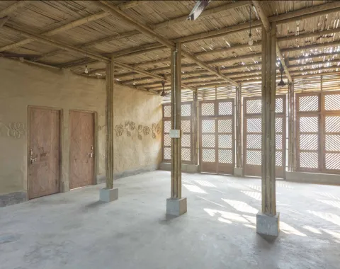 Alt-Text: Innenansicht eines nachhaltig gebauten Raumes mit Bambusstrukturen, Lehmwänden und Holztüren. Sonnenlicht fällt durch ein Gittermuster an den Fenstern und erzeugt ein warmes Ambiente.