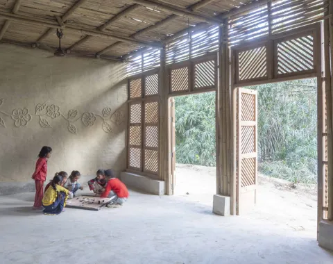 Einige Kinder spielen auf dem Boden eines hellen, nachhaltig gebauten Raumes mit großen Holztüren und dekorativen Wänden.