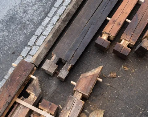 Holzbalken und Bauteile liegen auf einem gepflasterten Boden neben einem Bordstein. Die Materialien sind teilweise bearbeitet und bereit für den Einsatz in einem Bauprojekt. Das Bild zeigt den Fokus auf nachhaltige Baupraktiken und Wiederverwendung von Materialien.
