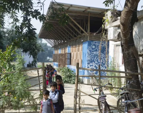 Eine Gruppe von Kindern geht einen Weg entlang, der zu einem nachhaltig gebauten Gebäude mit blauen Wänden und einem großen Dach führt. Im Vordergrund sind Bäume und ein Zaun zu sehen. Die Szene vermittelt eine ländliche, sonnige Atmosphäre.