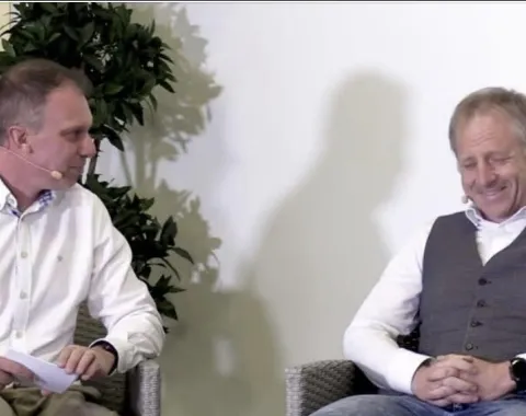 Zwei Männer sitzen auf einer Bühne und unterhalten sich. Beide tragen Mikrofone. Links sitzt ein Mann in einem weißen Hemd, rechts ein Mann in einem grauen Pullunder. Im Hintergrund ist eine Pflanze zu sehen.