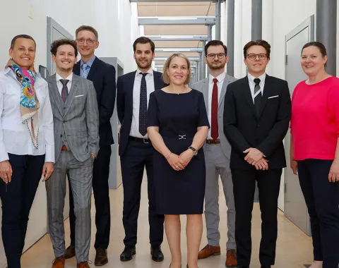 Eine Gruppe von acht Personen steht in einem Flur der Universität Liechtenstein. Sie tragen formelle Kleidung und lächeln in die Kamera. Die Umgebung ist modern und hell gestaltet.