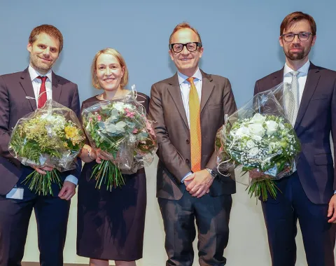 Vier Personen stehen nebeneinander auf einer Bühne. Drei von ihnen halten Blumensträuße in den Händen. Alle tragen formelle Kleidung und lächeln in die Kamera.