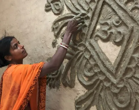 Eine Frau in einem orangefarbenen Sari verziert eine Wand mit traditionellen Tonmustern. Sie arbeitet konzentriert an einem kunstvollen Relief.