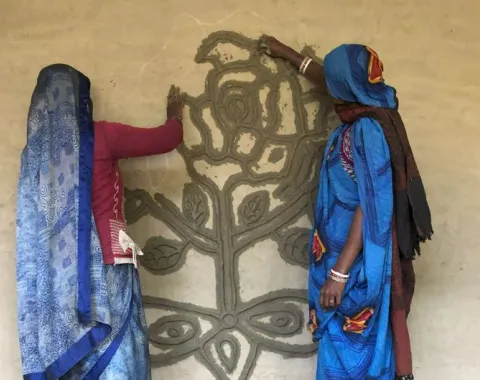 Zwei Frauen in traditionellen indischen Kleidern verzieren eine Wand mit einem kunstvollen Blumenmuster. Sie stehen auf einer Holzbank und arbeiten gemeinsam an der Gestaltung.