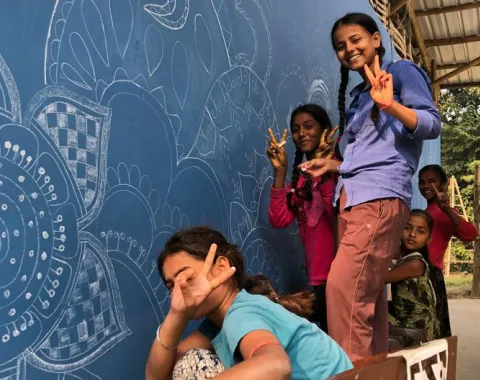 Kinder posieren fröhlich vor einer blau bemalten Wand mit kunstvollen weißen Mustern. Sie zeigen mit den Fingern das Friedenszeichen. Die Szene vermittelt Freude und Gemeinschaftsgefühl.