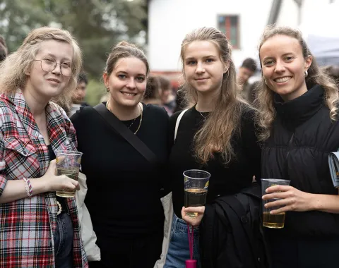 Vier junge Frauen stehen lächelnd zusammen auf einem Sommerfest der Universität Liechtenstein. Sie halten Getränke in der Hand und sind von anderen Feiernden umgeben. Im Hintergrund sind Bäume und ein Gebäude zu sehen.