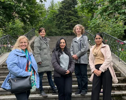 Fünf Personen stehen auf einer Treppe im Freien, umgeben von grünen Bäumen. Sie lächeln in die Kamera.