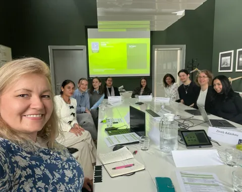 Eine Gruppe von sechs Personen steht lächelnd in einem Raum vor einer Wand mit gerahmten Bildern. Die Personen sind formell gekleidet und blicken in die Kamera.