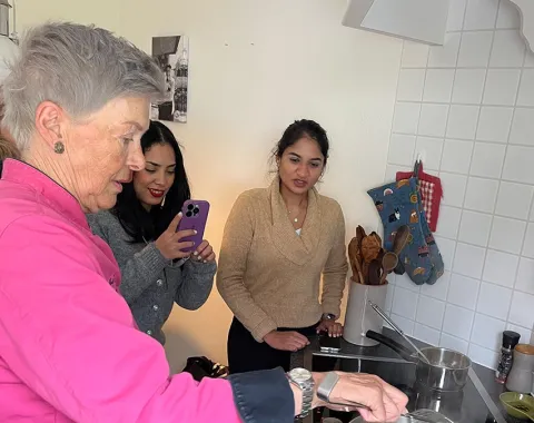 Eine Gruppe von Menschen steht in einer Küche. Eine Person in einem pinken Oberteil kocht an einem Herd, während zwei andere Personen zuschauen und eine von ihnen mit einem Smartphone fotografiert. Auf dem Herd steht ein Topf mit kochendem Wasser.
