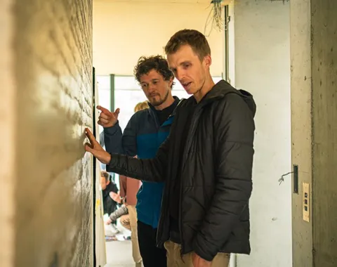 Zwei Männer betrachten und diskutieren eine Wand aus Stampflehm in einem Gebäude. Sie tragen Freizeitkleidung und befinden sich in einem hellen Raum.