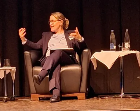 Eine Person sitzt auf einem Podium in einem Sessel und gestikuliert während einer Diskussion. Neben ihr stehen Tische mit Wasserflaschen und Gläsern. Die Szene findet in einem Konferenzsaal statt.