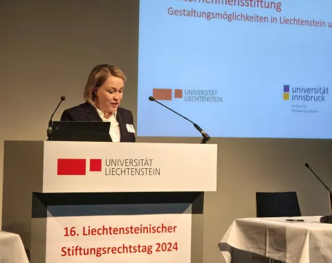 Eine Rednerin steht am Podium während des 16. Liechtensteinischen Stiftungsrechtstags 2024 an der Universität Liechtenstein. Im Hintergrund ist eine Präsentationsfolie mit Logos der Universität Liechtenstein und der Universität Innsbruck zu sehen.