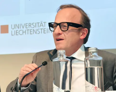 Ein Mann in Anzug und Brille spricht in ein Mikrofon auf einer Konferenz im Auditorium der Universität Liechtenstein. Vor ihm stehen Wasserflaschen. Im Hintergrund ist das Logo der Universität zu sehen.