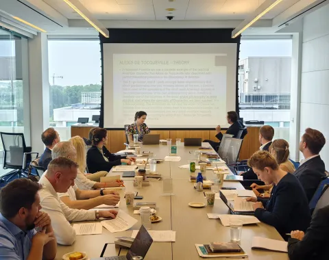 Ein Konferenzraum mit mehreren Personen, die an einem langen Tisch sitzen und einer Präsentation folgen. Im Hintergrund ist eine große Leinwand mit projiziertem Text zu sehen. Die Teilnehmer sind in eine Diskussion vertieft.