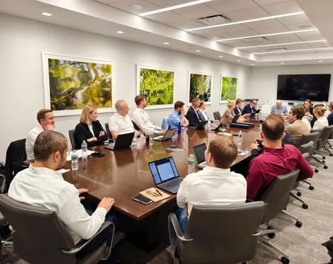 Eine Gruppe von Studierenden und Dozenten sitzt in einem modernen Konferenzraum an einem großen Tisch. Sie sind in eine Diskussion vertieft, umgeben von Laptops und Notizen. An den Wänden hängen Kunstwerke, und im Hintergrund ist ein großer Bildschirm zu sehen.