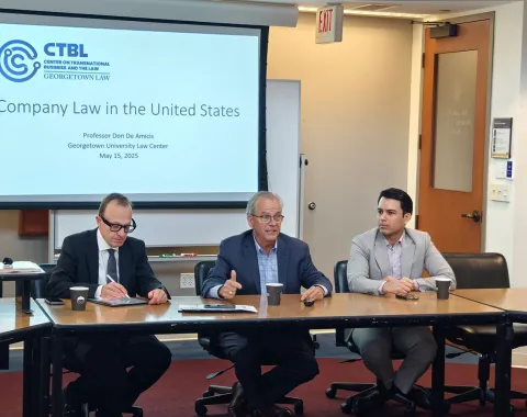Drei Personen sitzen an einem Konferenztisch in einem Seminarraum. Im Hintergrund ist eine Präsentationsfolie mit dem Titel "Company Law in the United States" zu sehen. Der Raum gehört zum Georgetown University Law Center.