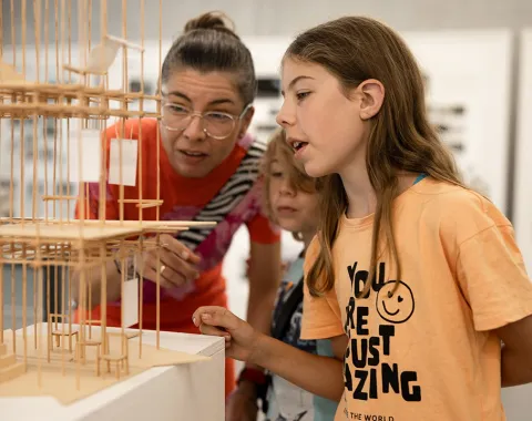 Eine Frau und zwei Kinder betrachten ein detailliertes Architekturmodell aus Holz. Die Szene findet in einem Ausstellungsraum statt, der Teil des Tags der offenen Tür an der Universität Liechtenstein ist.