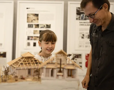 Ein Kind und ein Erwachsener betrachten ein detailliertes Architekturmodell eines Gebäudes. Im Hintergrund sind Informationsplakate zu sehen.