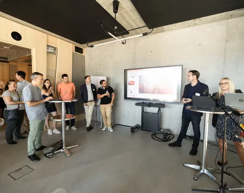 Eine Gruppe von Menschen steht in einem modernen Raum mit Betonwänden. Im Vordergrund sind Stehtische und ein großer Bildschirm an der Wand, auf dem eine Präsentation gezeigt wird. Eine Person spricht zu den Anwesenden, während andere aufmerksam zuhören. Der Raum wirkt hell und einladend.