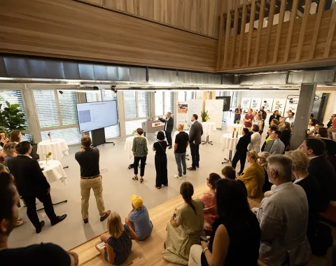 Eine Gruppe von Menschen versammelt sich in einem modernen, hellen Raum mit großen Fenstern. Im Vordergrund sitzen einige Personen auf Holzstufen. Im Hintergrund hält eine Person eine Präsentation vor einem Bildschirm. Der Raum ist mit Pflanzen und Stehtischen dekoriert.