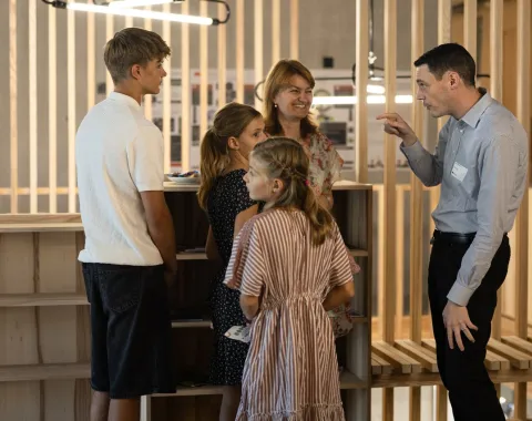 Eine Gruppe von Menschen steht in einem modernen Raum mit Holzelementen. Ein Mann spricht gestikulierend mit einer Frau und drei Kindern. Die Szene vermittelt eine freundliche und einladende Atmosphäre während des Tags der offenen Tür an der Universität Liechtenstein.