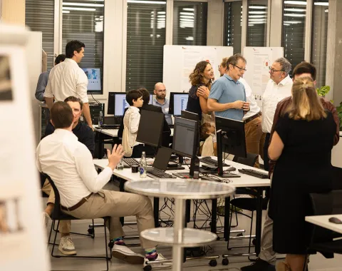 Eine Gruppe von Menschen unterhält sich in einem modernen Raum mit Computern und Informationspostern. Einige sitzen, während andere stehen und diskutieren. Die Atmosphäre wirkt lebhaft und interessiert, passend zu einem Tag der offenen Tür an der Universität.