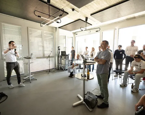 Eine Gruppe von Menschen steht in einem modernen, hellen Raum. Ein Mann hält eine Präsentation vor einem Publikum, das aufmerksam zuhört. Der Raum hat große Fenster und moderne Beleuchtung.