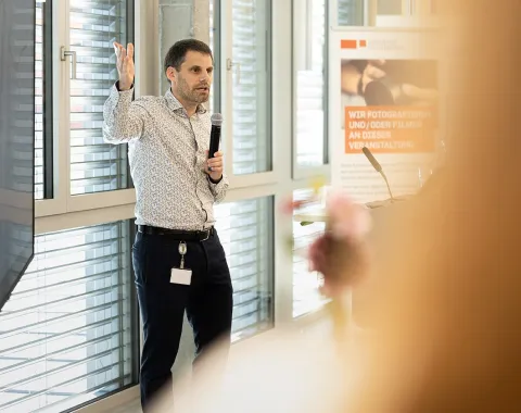 Eine Person hält eine Präsentation in einem modernen Raum mit großen Fenstern. Sie spricht in ein Mikrofon und gestikuliert mit der Hand. Im Hintergrund ist ein Plakat mit Informationen sichtbar.
