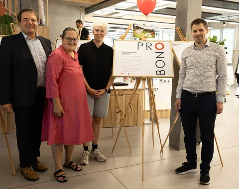 Vier Personen stehen in einem modernen Raum neben einer Ausstellungstafel mit der Aufschrift "Pro Bono". Im Hintergrund sind weitere Ausstellungstafeln und ein roter Luftballon zu sehen. Die Szene wirkt freundlich und einladend.