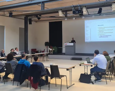 Eine Gruppe von etwa 20 Personen sitzt in einem Auditorium der Universität Liechtenstein, während ein Vortragender an einem Rednerpult spricht. Auf einer großen Leinwand ist eine Präsentation mit dem Titel "Wie weiter" zu sehen.