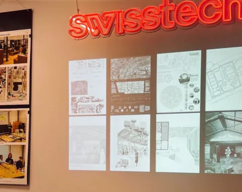 Ein Ausstellungstisch mit offenen Büchern steht vor einer Wand, auf der Architekturzeichnungen und Pläne projiziert sind. Links sind Fotos von Designprojekten zu sehen. Oben leuchtet ein "swisstech" Schild in roter Schrift.
