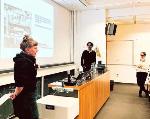 Eine Präsentation in einem Universitätsseminarraum: Drei Personen stehen vor einer Leinwand, auf der eine Folie projiziert wird. Eine Person spricht, während die anderen zuhören. Der Raum ist mit Tischen und einer Tafel ausgestattet.