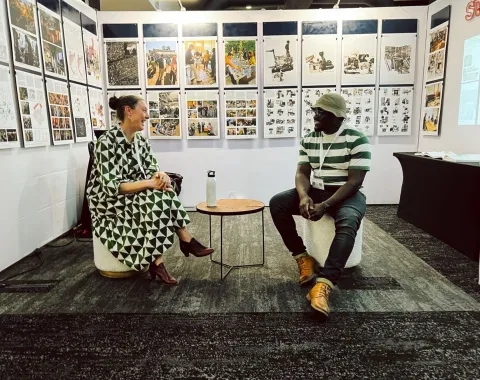 Zwei Personen sitzen in einer Ausstellung vor einer Wand mit zahlreichen Bildern und Texten. Sie unterhalten sich entspannt auf Hockern. Die Ausstellung thematisiert urbanes Design und soziale Realitäten in Afrika.