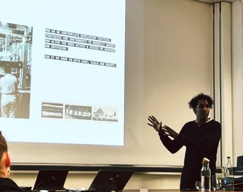 Ein Vortragender steht vor einer Projektionsleinwand und präsentiert Inhalte zu urbanem Design. Auf der Leinwand sind Text und Bilder zu sehen. Im Vordergrund sitzen Zuhörer. Der Raum wirkt modern und technisch ausgestattet.