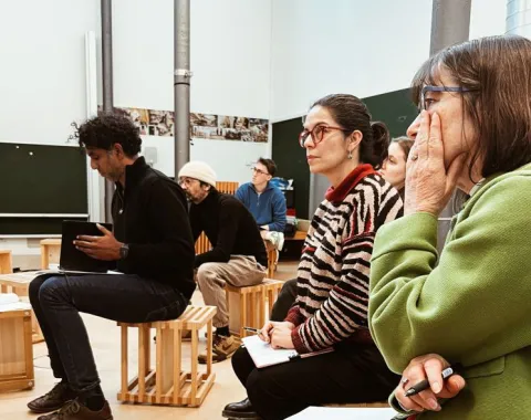 Eine Gruppe von Personen sitzt aufmerksam in einem Seminarraum. Sie sind auf Holzhockern platziert und scheinen einer Präsentation oder Diskussion zu folgen. Im Hintergrund sind Tafeln und Poster zu sehen. Die Szene vermittelt eine konzentrierte Lernatmosphäre.