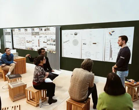 Eine Gruppe von Studierenden sitzt auf Hockern in einem Seminarraum und diskutiert vor einer Wand mit Architekturplänen und Diagrammen. Ein Präsentierender steht rechts und erklärt die Inhalte.