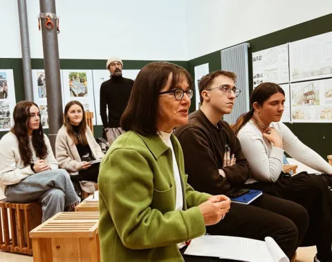 Eine Gruppe von Studierenden und Dozenten sitzt in einem Seminarraum und hört aufmerksam zu. Im Hintergrund sind Präsentationstafeln mit städtischen Entwürfen zu sehen.