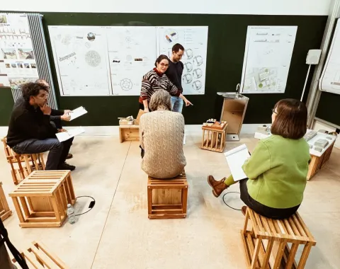 Eine Gruppe von sechs Personen sitzt auf Holzkisten in einem Raum mit Plakaten an der Wand. Zwei Personen stehen und diskutieren, während die anderen Notizen machen. Der Raum wirkt wie ein Workshop oder eine Präsentation.