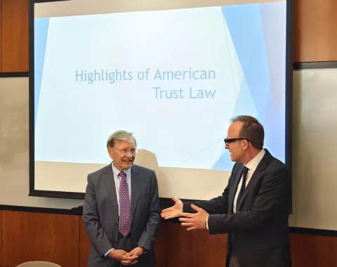 Zwei Männer in Anzügen stehen vor einer Präsentation mit dem Titel "Highlights of American Trust Law". Einer der Männer spricht gestikulierend. Im Hintergrund ist ein Projektionsbildschirm zu sehen.