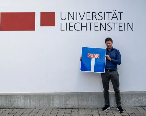Ein Mann steht vor einer Wand mit dem Logo der Universität Liechtenstein und hält ein Verkehrsschild mit der Aufschrift „Krieg“ in einer Sackgasse.