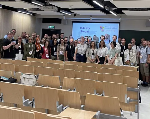 Eine Gruppe von Menschen steht in einem Hörsaal vor einer Präsentationsleinwand. Die Leinwand zeigt den Titel "Industry Forum". Die Personen lächeln in die Kamera. Im Vordergrund sind leere Sitzreihen zu sehen.